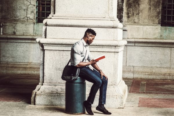 African American College Student studying in New York. Wearing gray shirt, jeans, cloth shoes, carrying shoulder leather bag, a black man sitting on metal pillar on street, relaxing, reading red book.