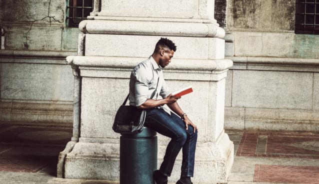 African American College Student studying in New York. Wearing gray shirt, jeans, cloth shoes, carrying shoulder leather bag, a black man sitting on metal pillar on street, relaxing, reading red book.